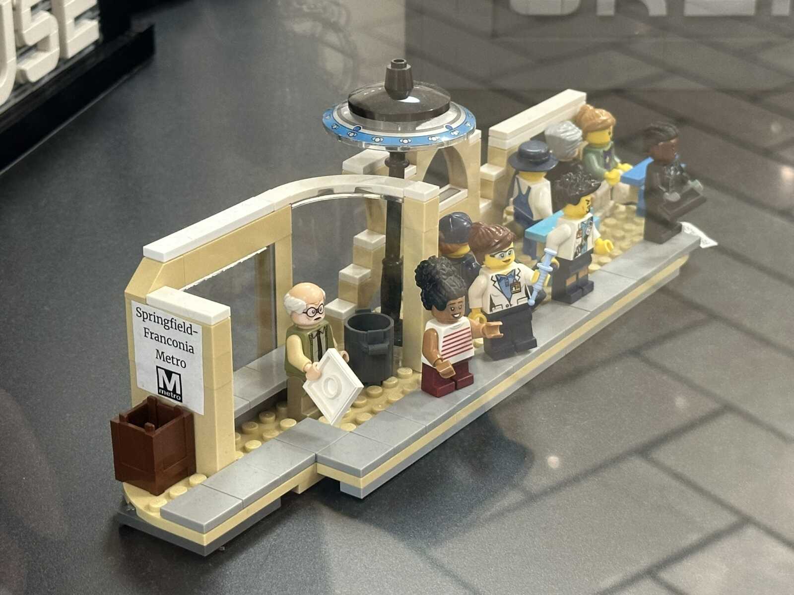 LEGO metro station scene with diverse minifigures waiting on a tan platform, sign reads Springfield-Franconia Metro, futuristic blue ring overhead.