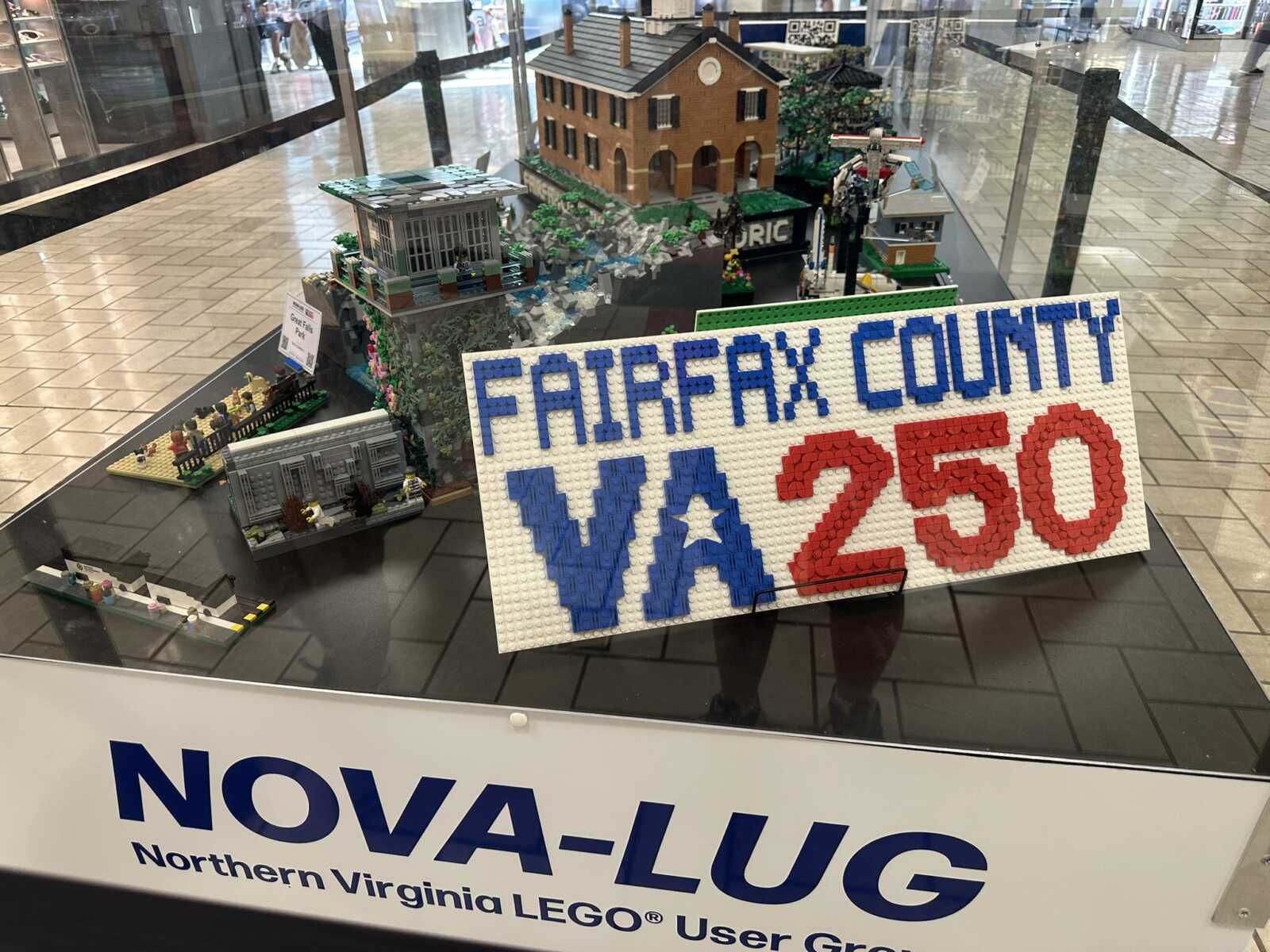 LEGO display showing a town scene with a brick house and miniature structures, and a large sign reading 'FAIRFAX COUNTY VA 250' in blue and red bricks