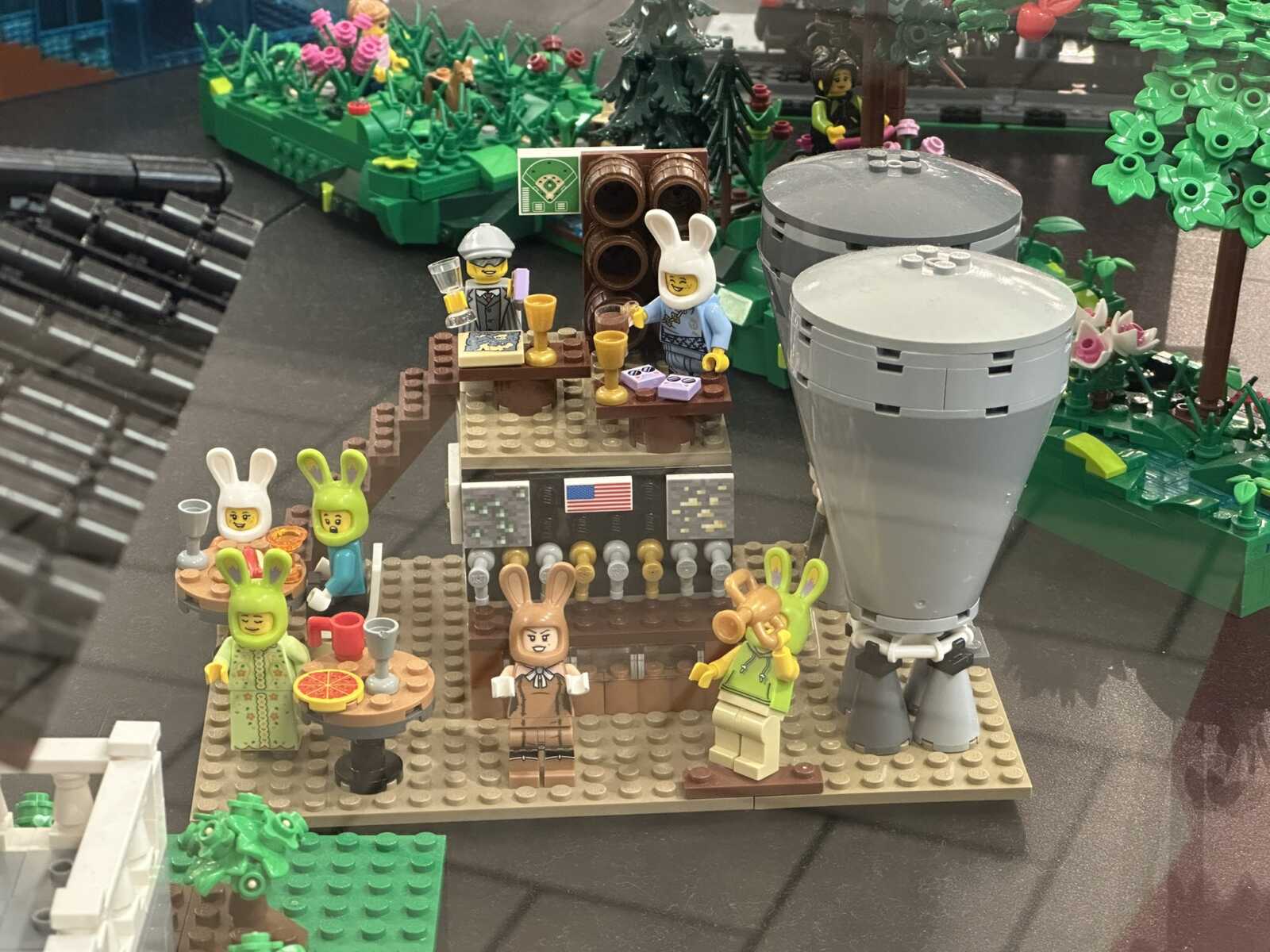 LEGO scene: four bunny minifigures around a small table with drinks outdoors, beside a tall gray cylindrical structure amid trees and flowers.