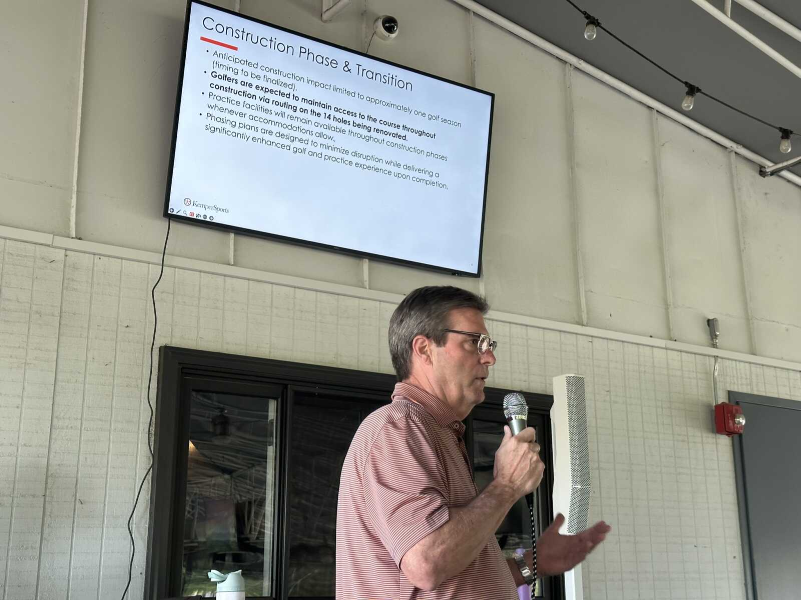 Man speaking into a microphone in front of a projected slide that reads 'Construction Phase & Transition' in an indoor setting