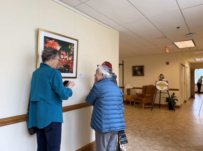 Two elderly people examine a framed painting on a wall in a bright hallway.