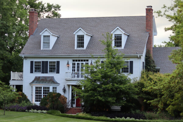 White colonial-style house with dormer windows, black shutters, a red front door, brick chimney, and lush landscaping in front.