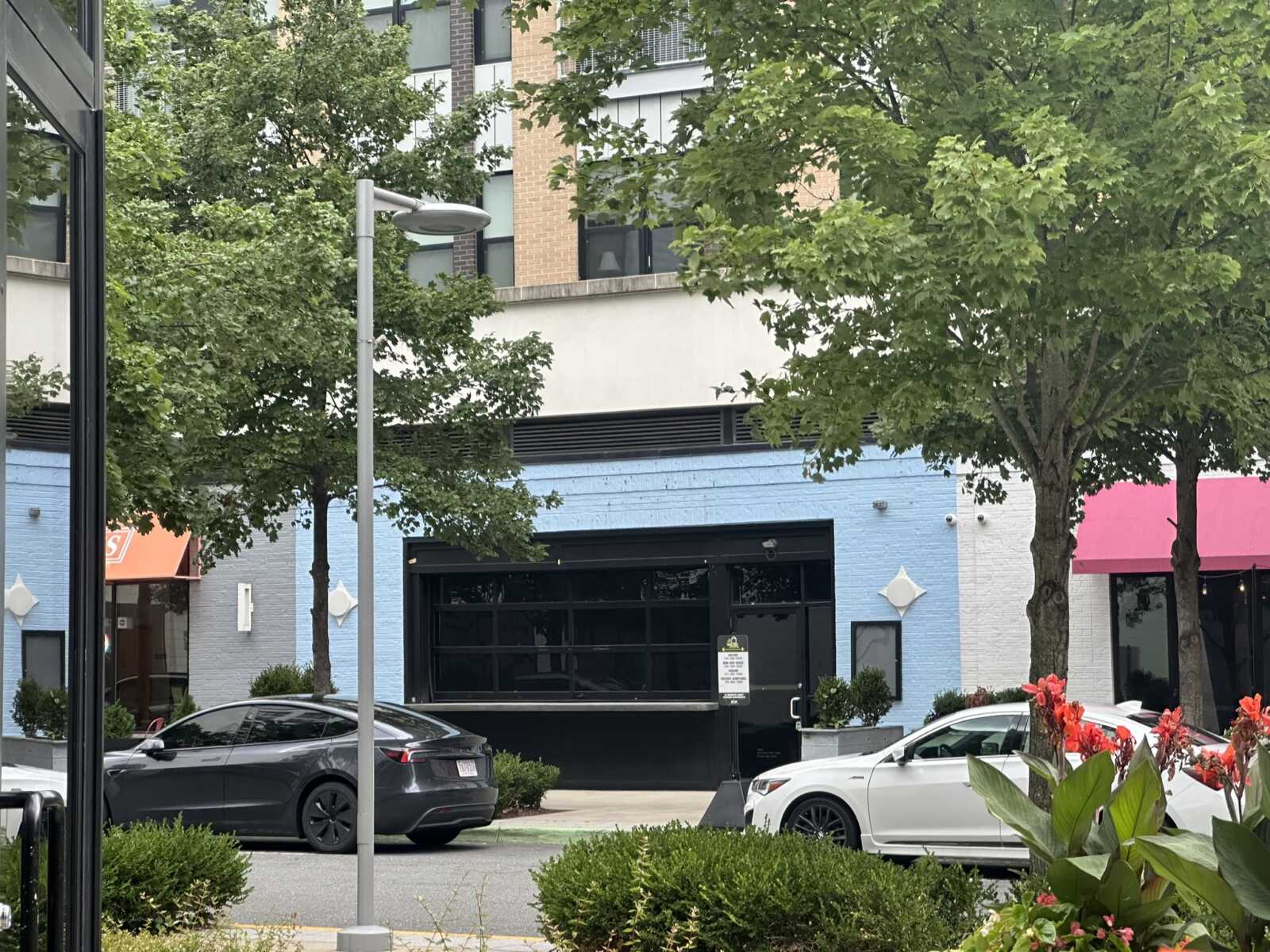 City street view with trees, a blue storefront wall, a large black garage door, and parked cars in the foreground.