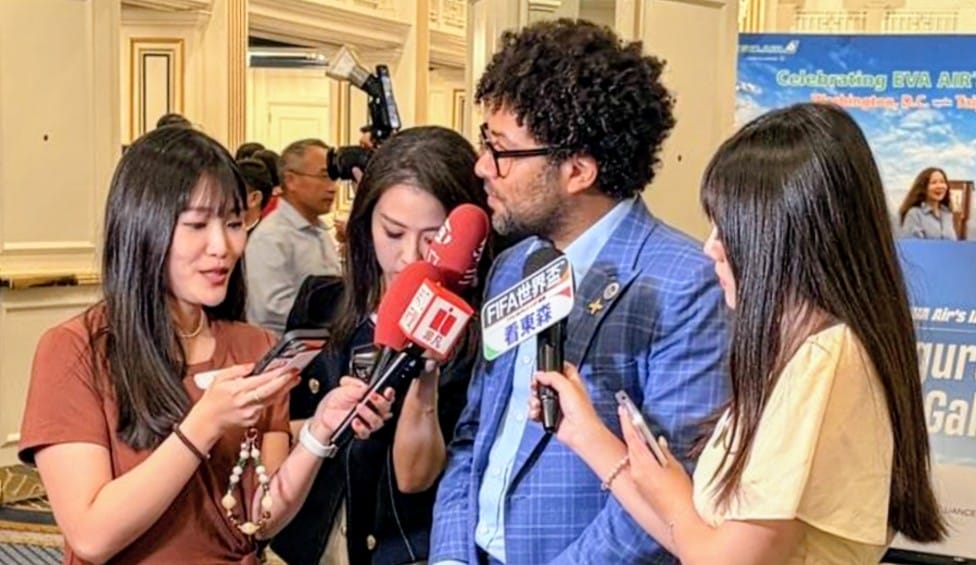 Paul Bobson discusses Dulles International Airport with media in South Korean (via MWAA) | FFXnow Man in a blue blazer is interviewed by reporters holding red and branded microphones at a press event inside a hotel hall.