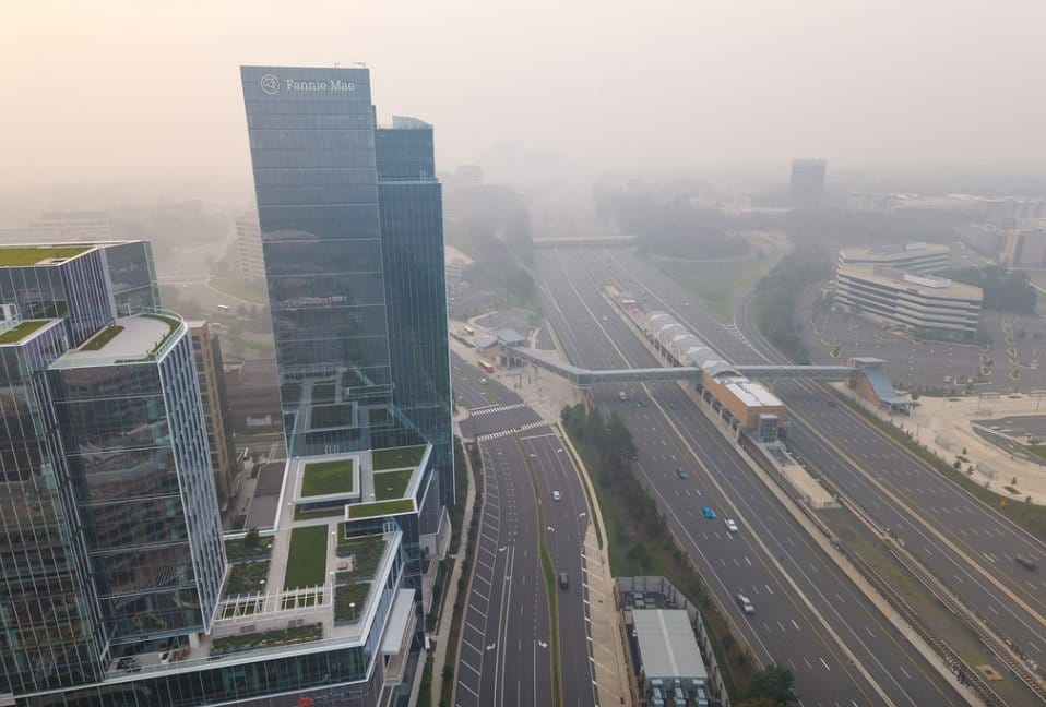 Aerial view of glass office towers, including a tall Fannie Mae building, with a multi-lane highway and an elevated pedestrian bridge in a hazy cityscape.
