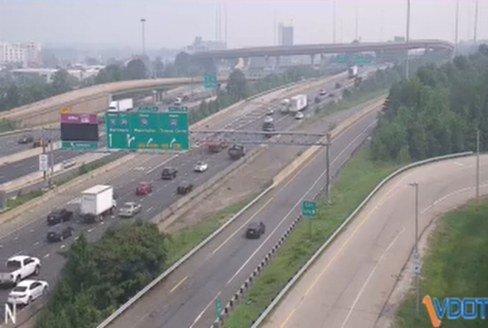 Aerial view of a multi-lane highway with vehicles moving in both directions and green overhead exit signs, hazy city in the background.