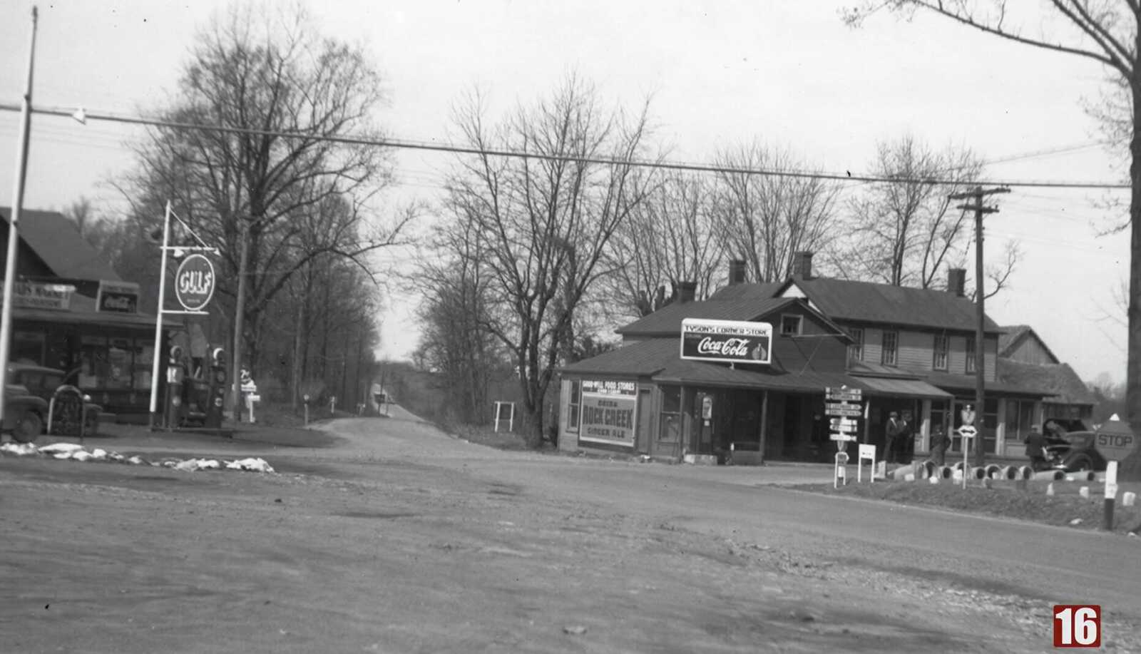 Gas station and corner store at the intersection of Leesburg Pike and Dolley Madison Blvd in the 1930s (via Fairfax County Channel 16) | FFXnow Historic small-town street scene with gas station and storefronts, Coca-Cola sign, Gulf sign, and leafless trees on a dirt road edge.
