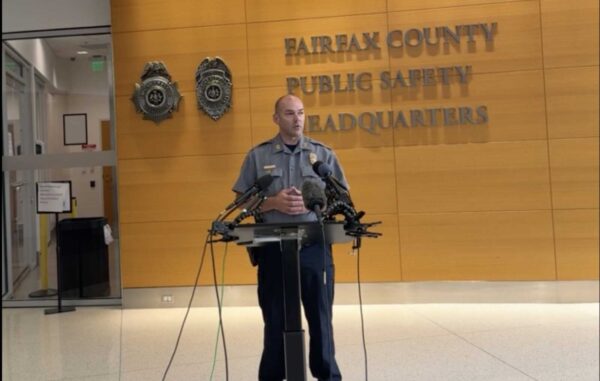Police officer stands at a podium with multiple microphones in Fairfax County Public Safety Headquarters lobby.