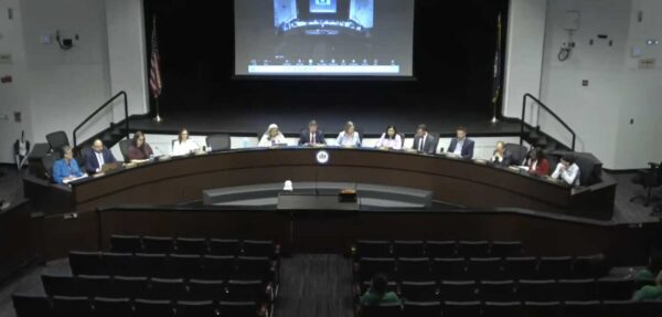 Panel of officials seated at a long curved desk on a stage during a formal meeting, projector screen overhead, audience in foreground.