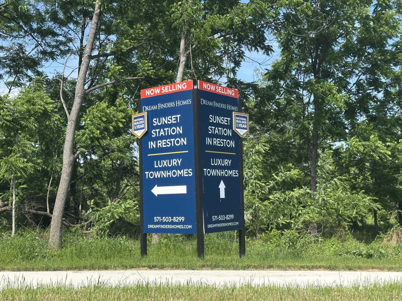 Two blue real estate signs read Sunset Station in Reston for luxury townhomes, with arrows left and up, under a Now Selling banner by trees.