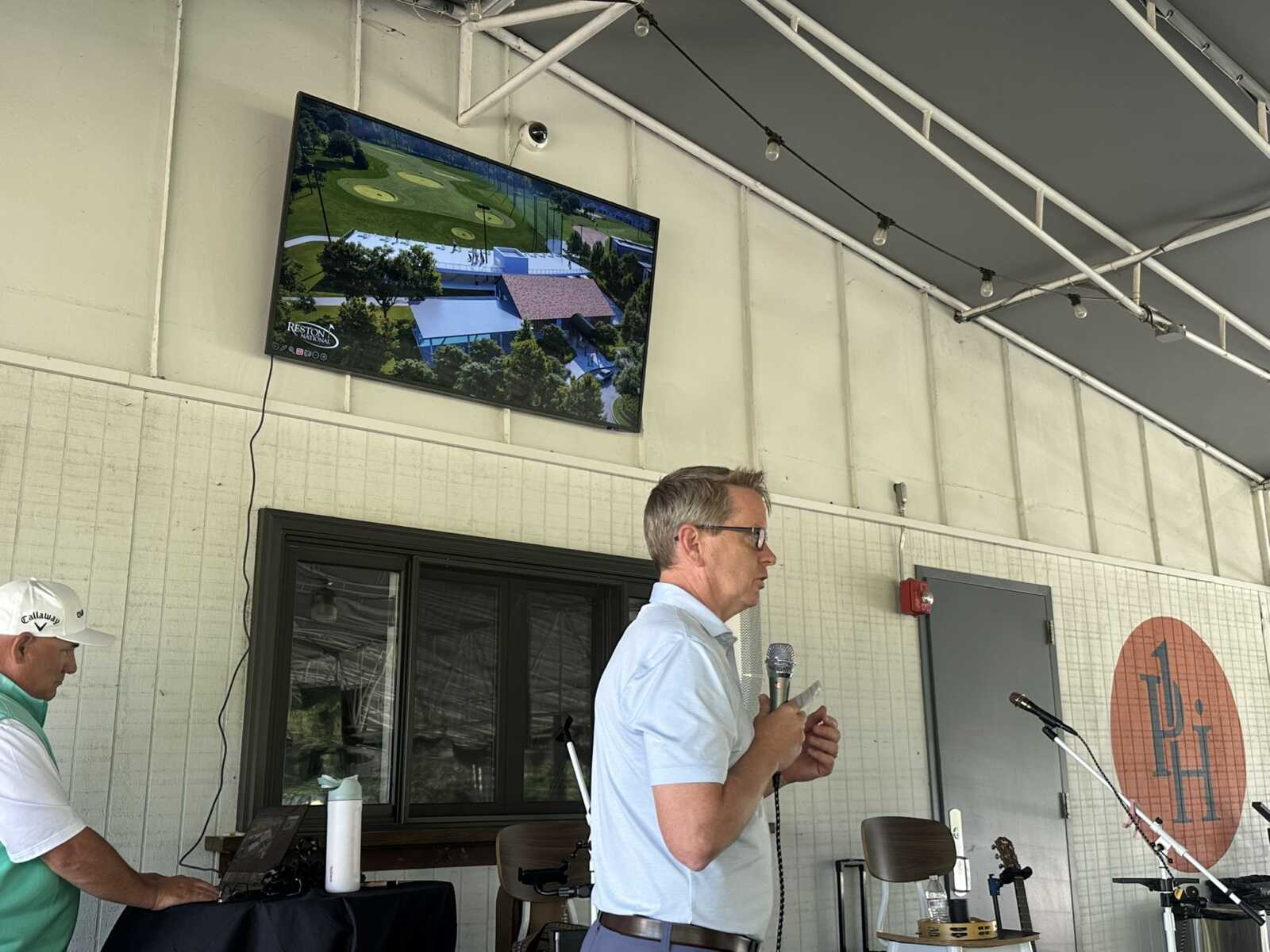 Man in a light blue shirt speaks into a handheld microphone at a golf event on a covered patio, with a TV showing a golf course behind him