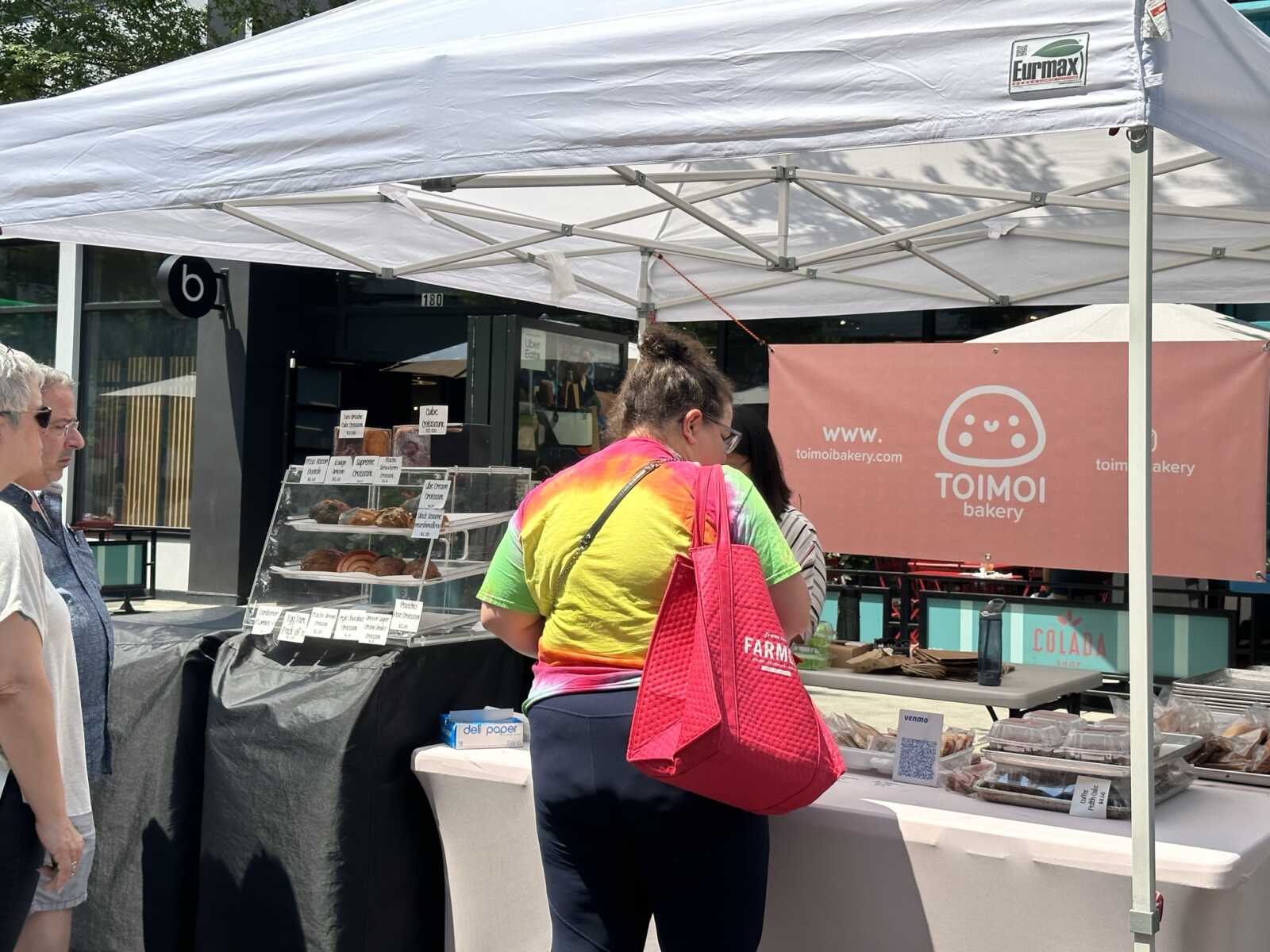 Person in a colorful shirt with a bright pink bag buys pastries at an outdoor bakery stall under a white canopy.