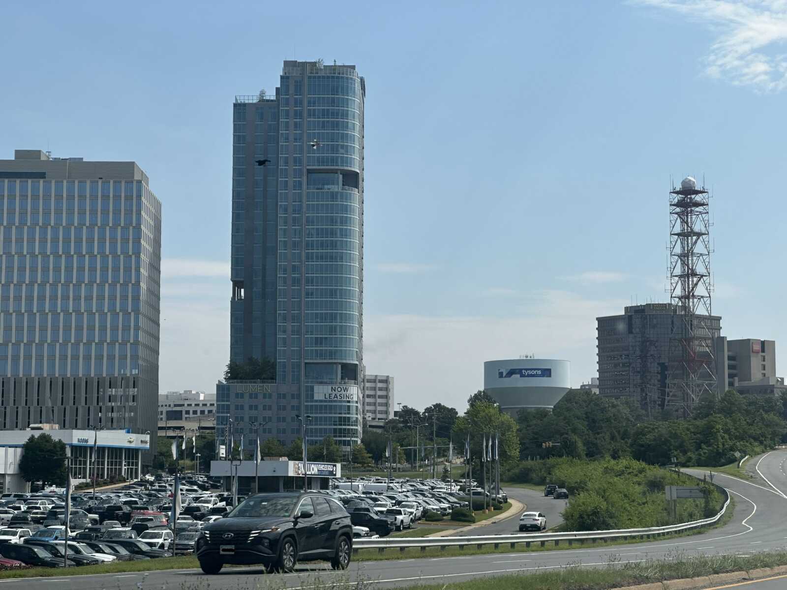 The Tysons water tower and Lumen Apartments next to the Route 7 and Route 123 interchange (staff photo by Angela Woolsey) | FFXnow City skyline with a tall glass office tower, surrounding buildings, a large parking lot, and a communications tower against a clear blue sky.