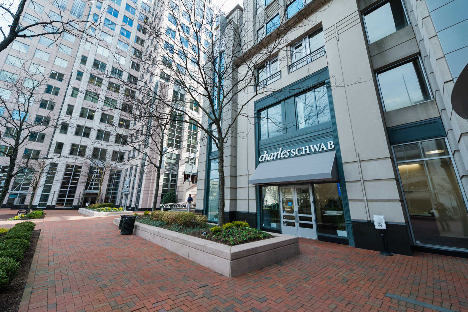 Charles Schwab storefront in a modern high-rise complex with a brick-paved plaza and leafless trees in the foreground.