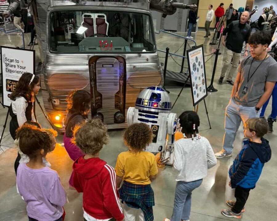 Children gather around a life-size R2-D2 droid beside a silver retro vehicle on a museum display, with onlookers in the background and a sign nearby.