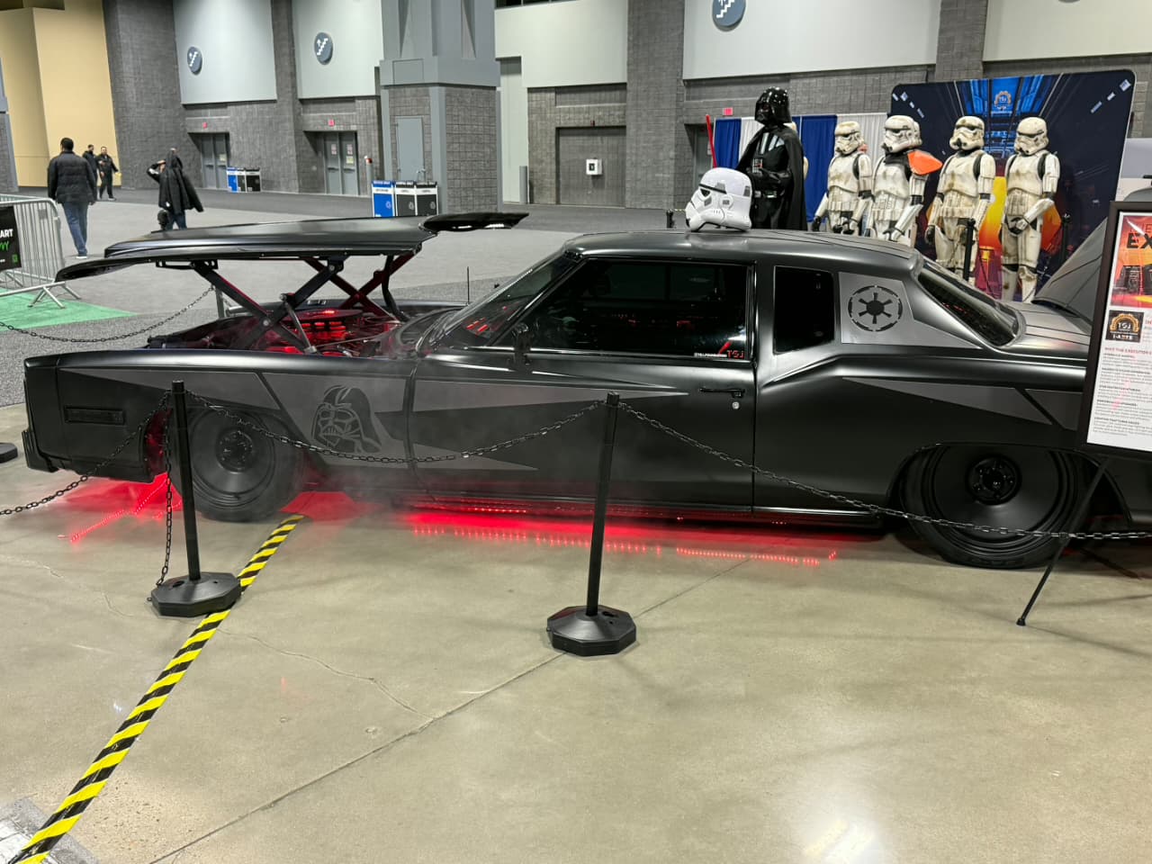 Black customized car with red underglow and a Darth Vader helmet on the roof, displaying Imperial logos at a Star Wars exhibit.