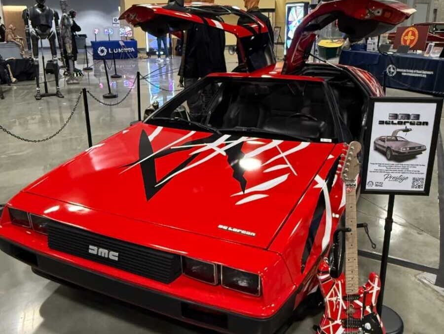 Front view of a red DeLorean-style car with gull-wing doors open, on display at an indoor car show with a guitar leaning nearby.
