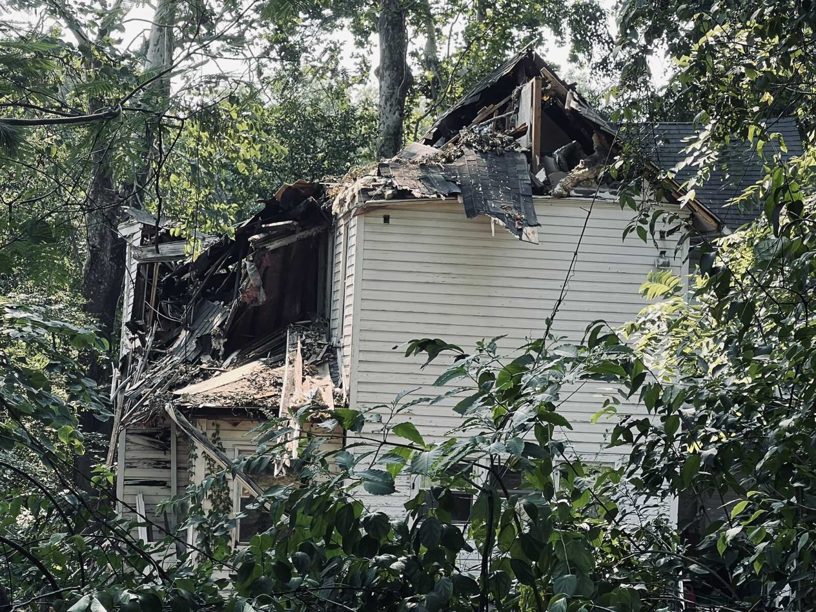 A collapsed, derelict house partially hidden by dense green foliage and trees in a wooded area, with broken roof and debris scattered around.
