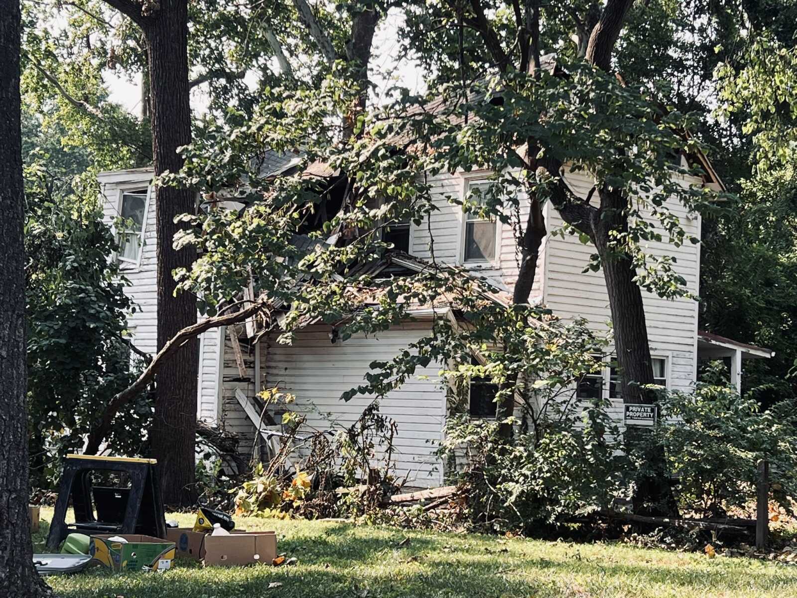 House partially crushed by a large fallen tree, with broken roof and scattered debris in the yard.
