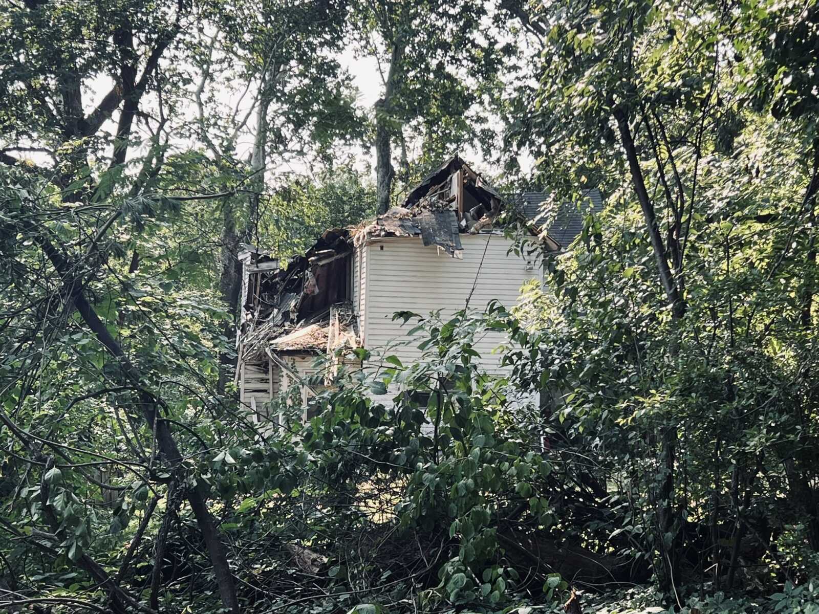 Partially collapsed two-story house overgrown by trees, with a damaged roof and exposed framing amid dense vegetation.