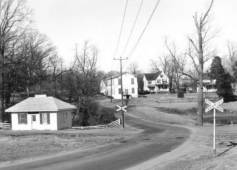 The Wiehle area of Reston in the 1950s (courtesy Reston Historic Trust and Museum) | FFXnow Rural road at a railroad crossing with a small white building on the left and houses in the background under bare trees.