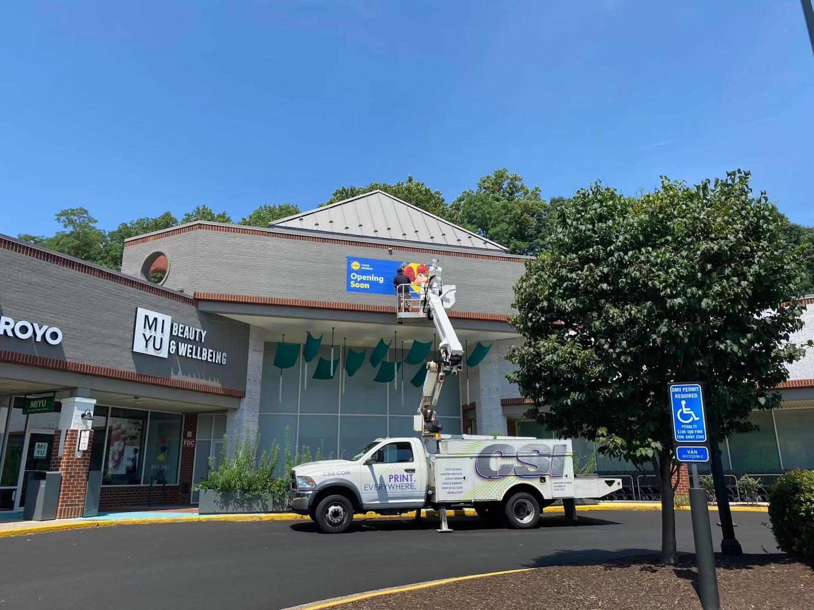 Retail storefront with 'MIYU Beauty & Wellbeing' signage and a 'Opening Soon' banner being installed above the entrance by a worker in a bucket truck.