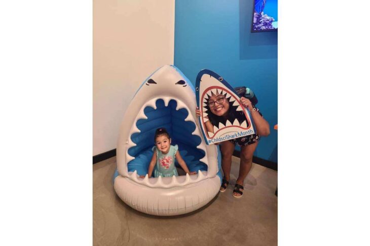 Young girl sits inside a white-and-blue inflatable shark mouth; a smiling woman holds a shark-shaped sign beside her in a colorful room.