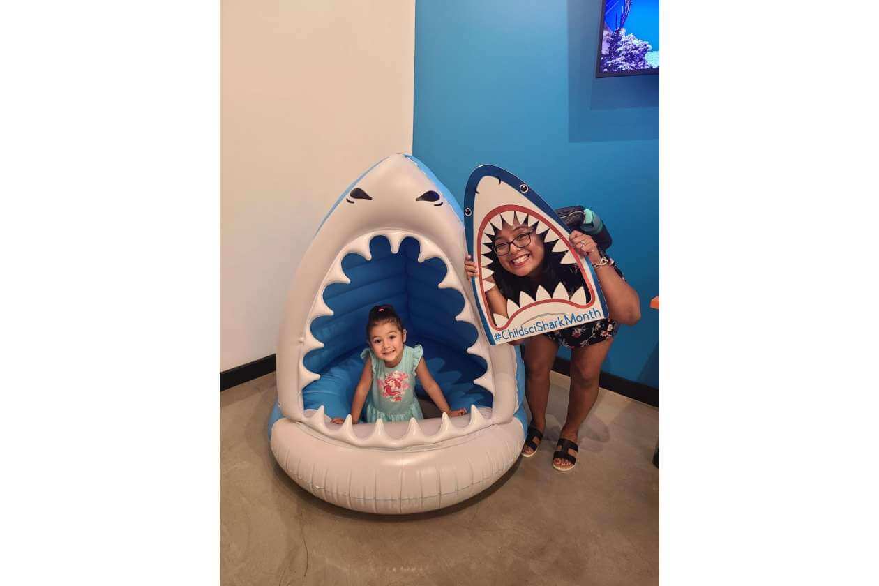 Young girl sits inside a white-and-blue inflatable shark mouth; a smiling woman holds a shark-shaped sign beside her in a colorful room.