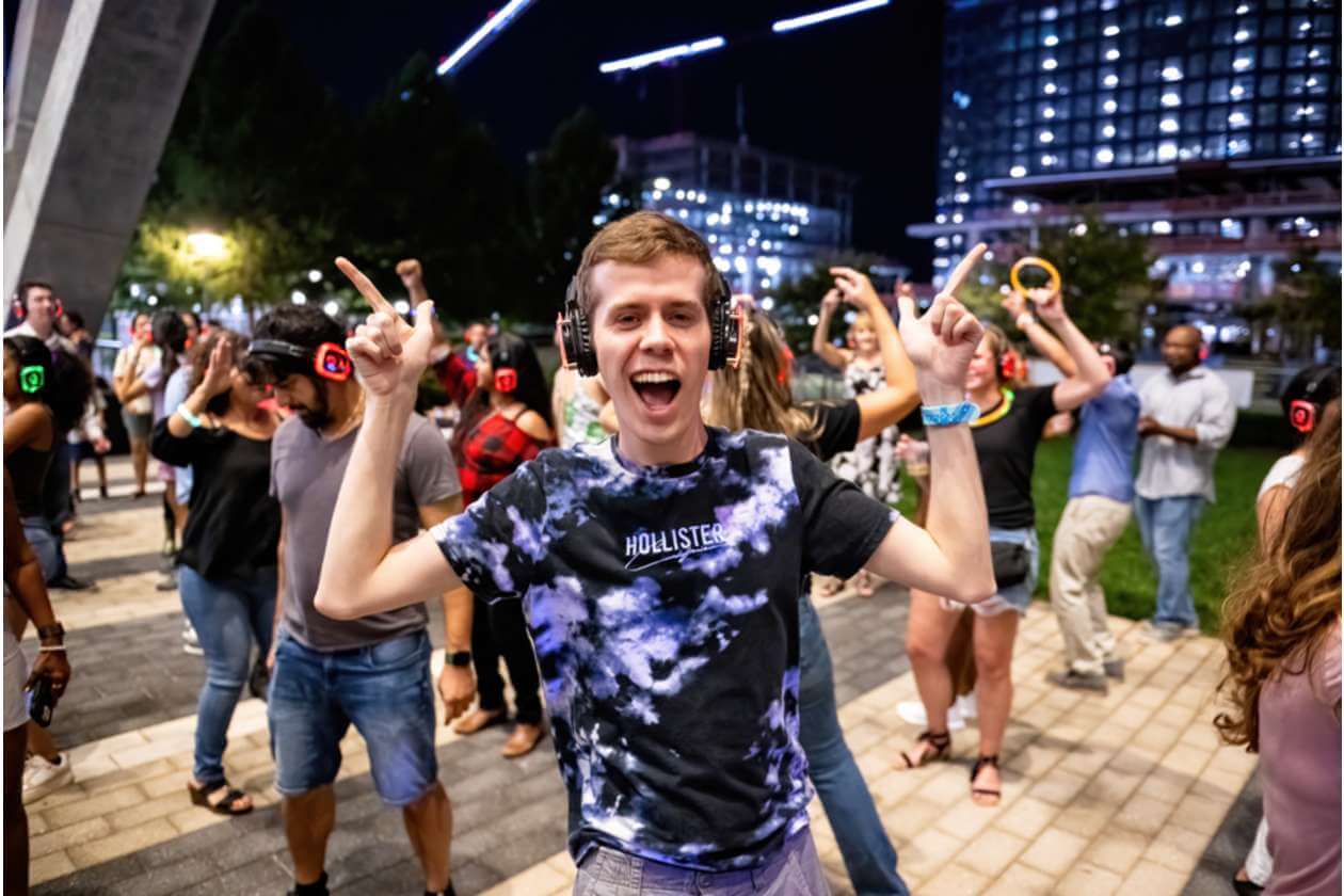 Young man wearing headphones dances at a silent disco outdoors at night with a cheering crowd behind him.