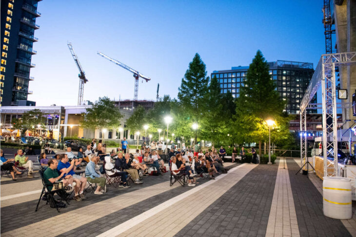 Audience seated on folding chairs at an outdoor evening event in a city plaza, stage on the right with lights.