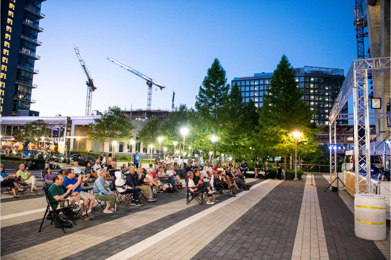Audience seated on folding chairs at an outdoor evening event in a city plaza, stage on the right with lights.