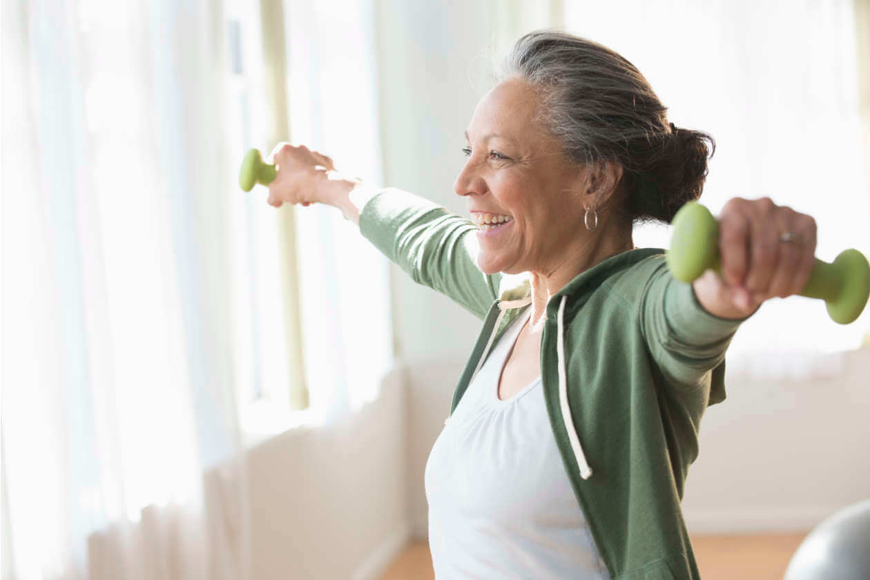 Older woman exercising with light green dumbbells, arms extended, smiling in a bright room wearing a green zip hoodie and white top.
