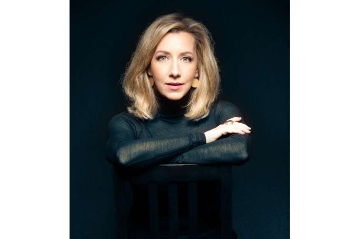 Portrait of a blonde woman in a black turtleneck resting her arms on the back of a chair against a dark background.