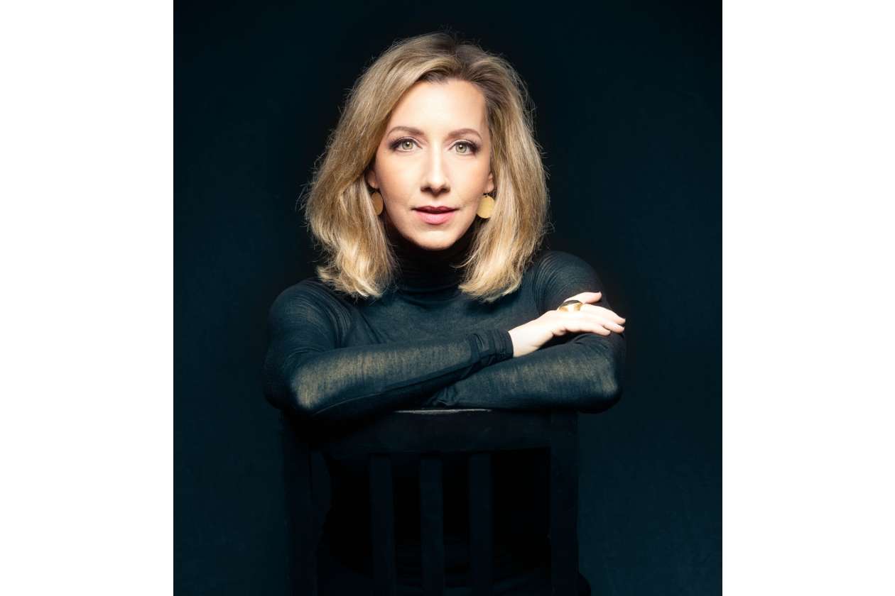 Portrait of a blonde woman in a black turtleneck resting her arms on the back of a chair against a dark background.