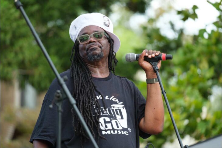 African American man wearing a white bucket hat and sunglasses sings into a handheld microphone on an outdoor stage; his shirt reads GO MUSIC.