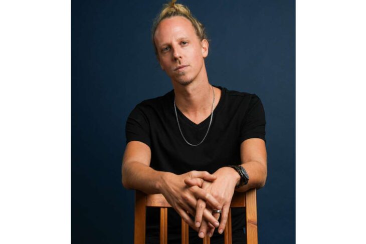 Portrait of a man with blond hair tied back, wearing a black T-shirt and a silver necklace, seated with hands clasped on a wooden chair against a dark blue background.
