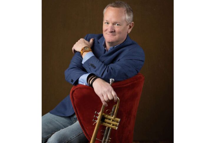 Portrait of a man in a blue blazer sitting on a red chair, resting a trumpet on his lap.
