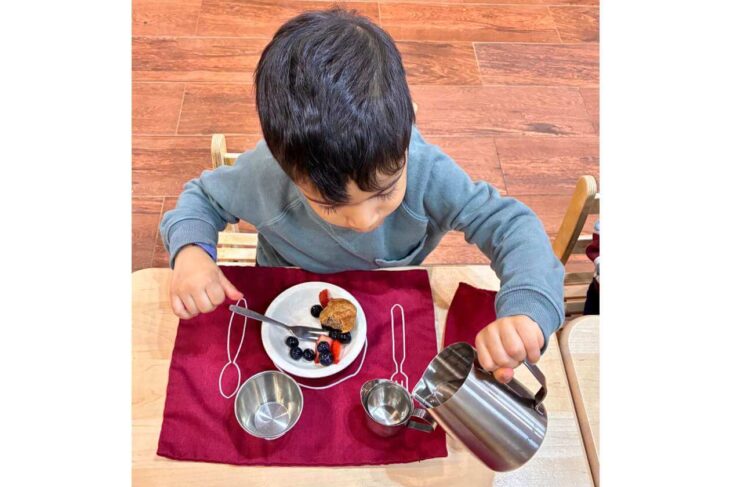 Young child at a table pours liquid from a metal pitcher into a cup beside a plate of berries and a pastry on a red placemat.