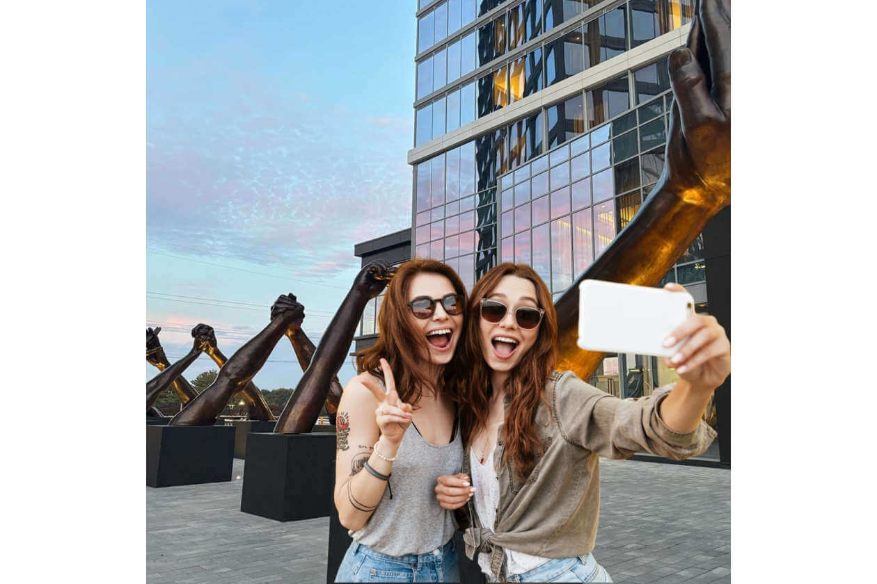 Two friends wearing sunglasses take a selfie with a smartphone in front of a glass-fronted building and dark sculptures outdoors on a sunny day.