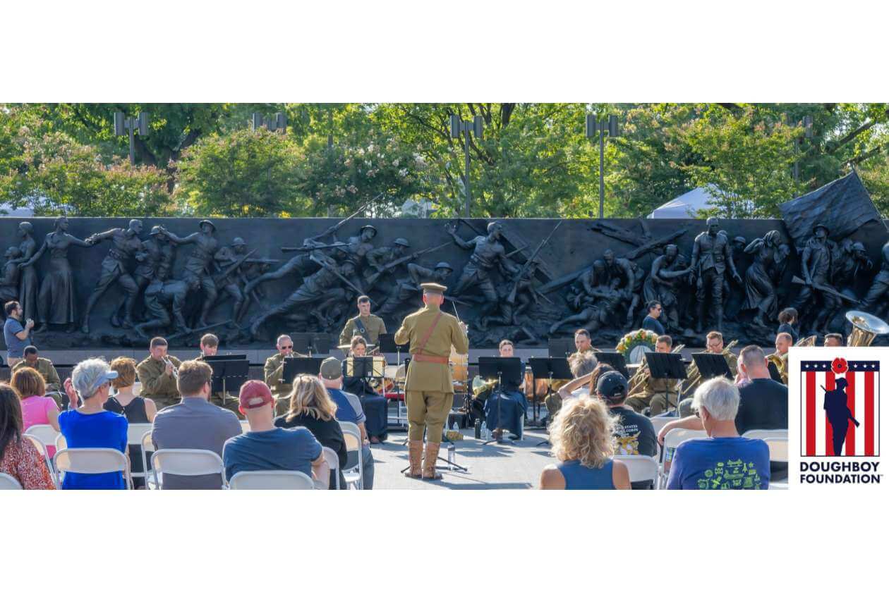 Military band performing outdoors with conductor in front, audience seated, large sculpted wall behind, Doughboy Foundation logo in corner.