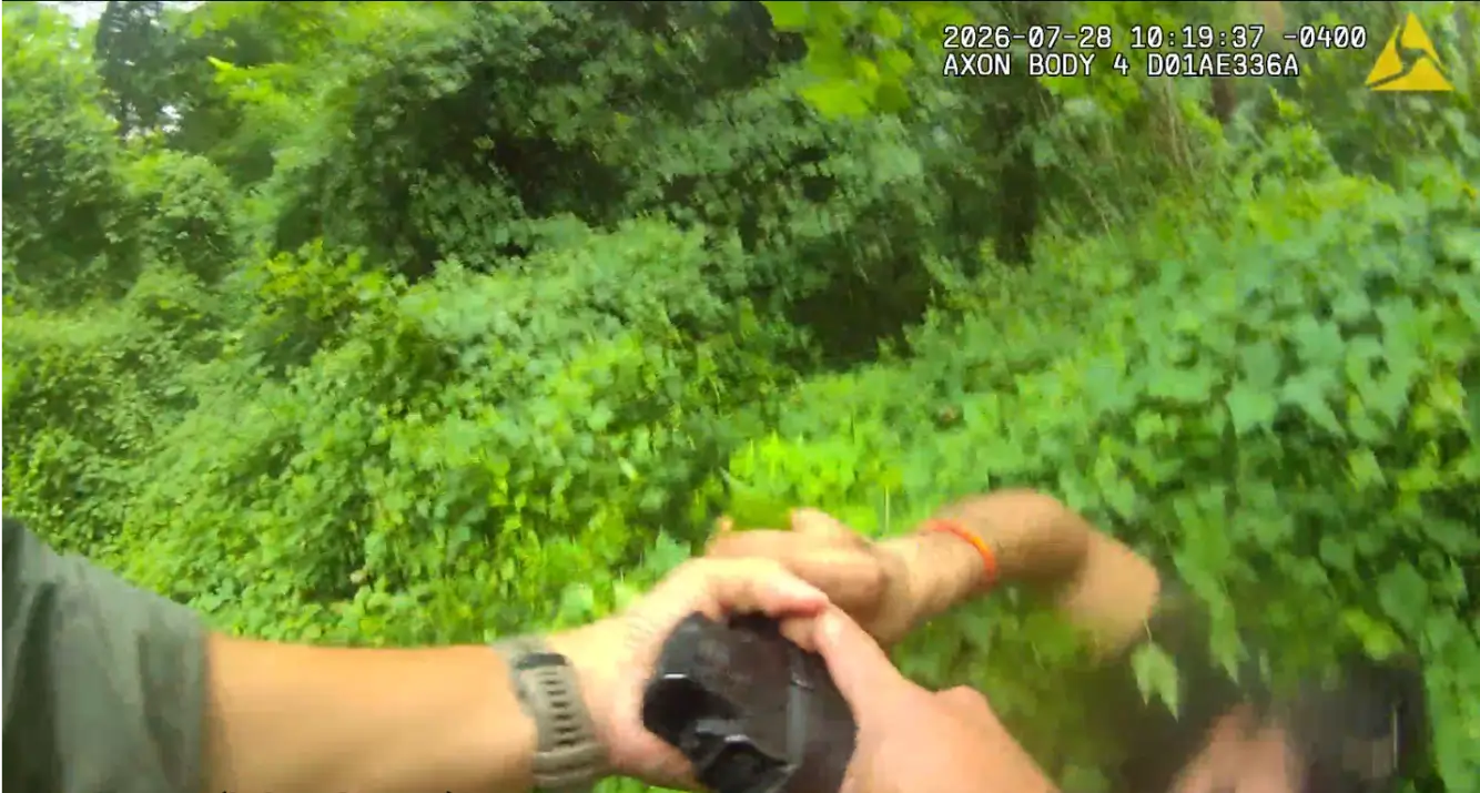 First-person view from a body camera in a dense green outdoor area; hands grip a black device in the foreground, timestamp shown.