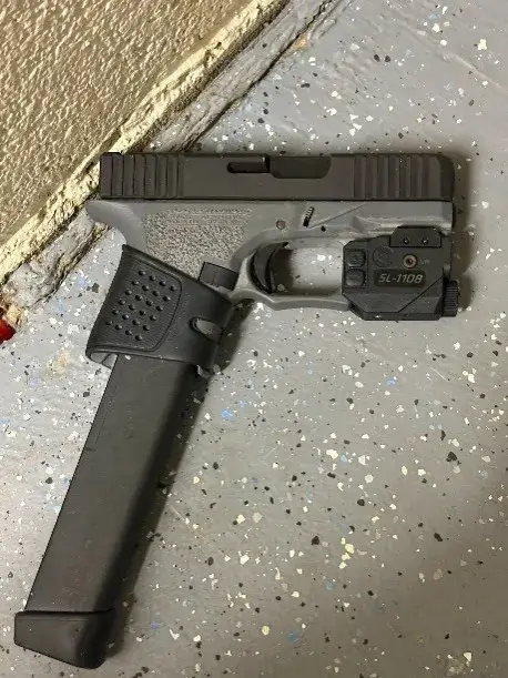 Semi-automatic pistol with an extended magazine and mounted optic resting on a speckled concrete floor by a wall edge.