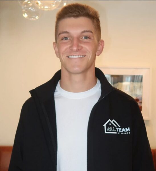 Smiling young man in a white shirt and black jacket with an All Team Exteriors logo, indoors.