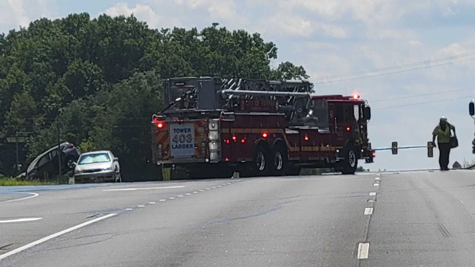 Fire truck with an extended ladder blocking a road; emergency lights on, a car off the roadway to the left, and a worker in a reflective vest nearby.