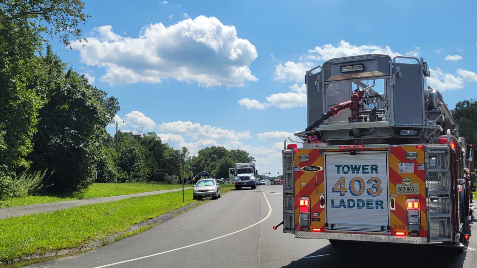 Fire truck with a tall ladder labeled 'Tower 403' occupying a sunny two-lane road by green grass and trees on the left.