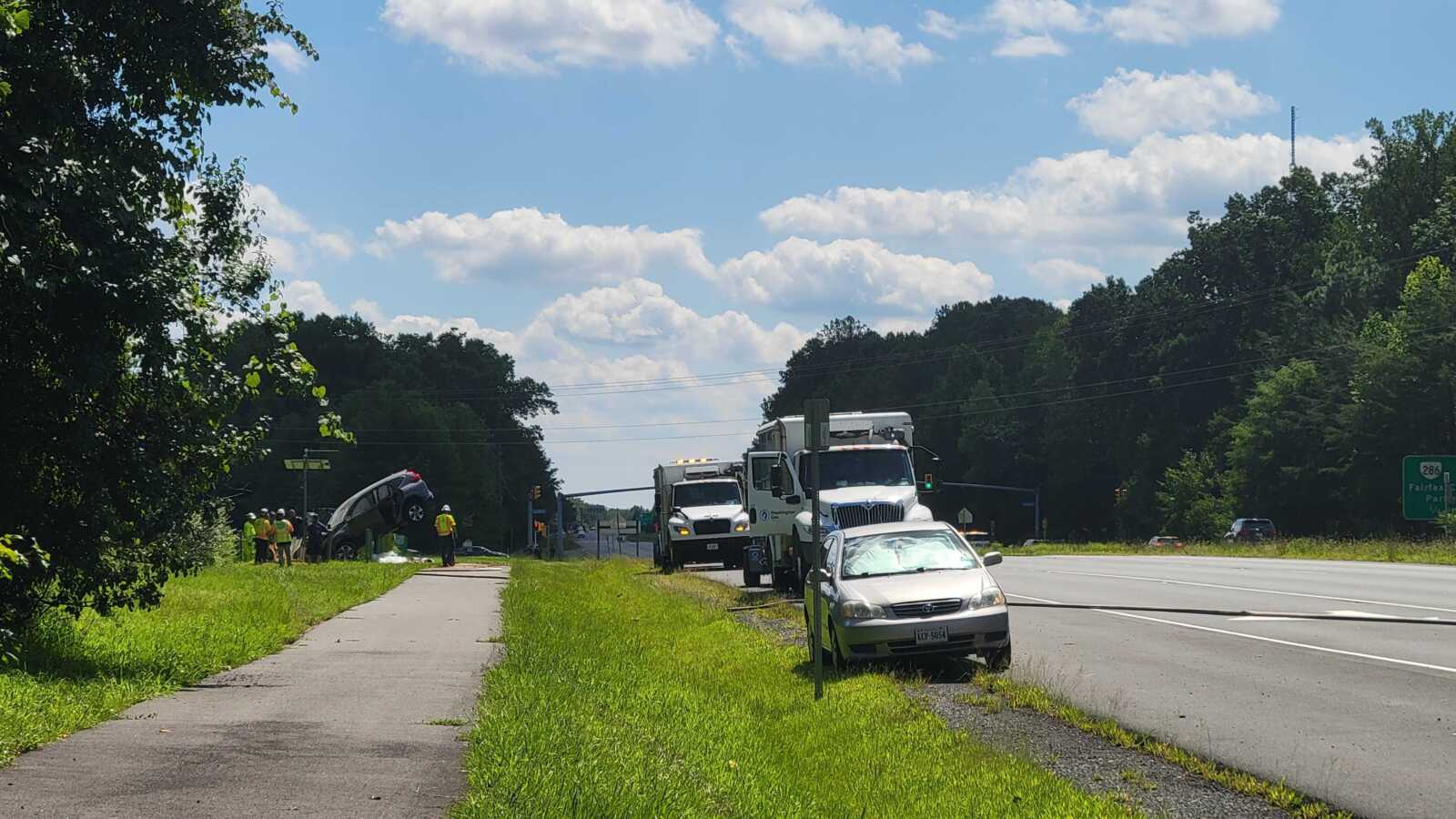 Emergency responders in yellow vests inspect a car crash on the highway shoulder near a grass verge beneath a clear sky.