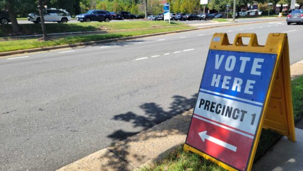 Election sign on a yellow stand along a sidewalk reads 'VOTE HERE' and 'PRECINCT 1' with a left arrow, directing voters to a polling location.