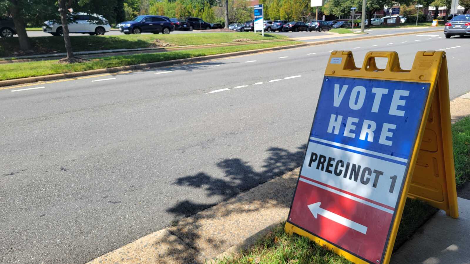 Election sign on a yellow stand along a sidewalk reads 'VOTE HERE' and 'PRECINCT 1' with a left arrow, directing voters to a polling location.