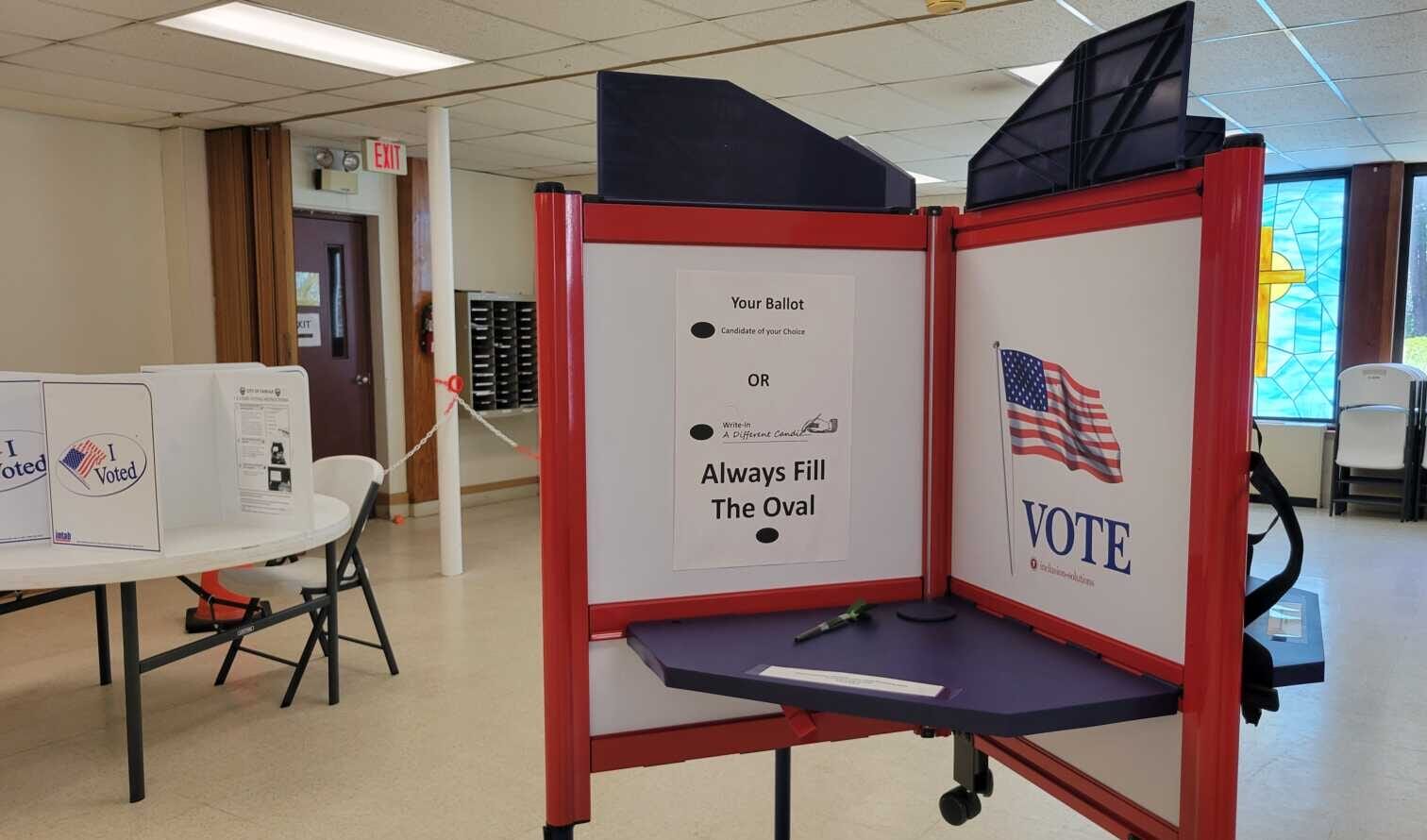 A voting booth with red panels, a blue work surface, and a sign reading 'Your Ballot' inside; an 'Always Fill The Oval' instruction is visible. A large poster with an American flag and the word VOTE is on the side panel in the background.