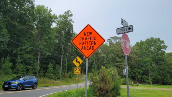Orange diamond sign reads 'New Traffic Pattern Ahead' with a winding road sign and 20 mph sign nearby by a rural road.